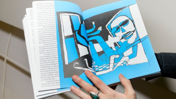 Eine Person
                                          hält ein aufgeschlagenes Buch in den Händen. Auf der rechten Doppelseite ist eine stilisierte Illustration in Blau, Schwarz
                                          und Weiß zu sehen: eine Figur sitzt vor einem Computerbildschirm. Die linken Seiten enthalten Fließtext. Die Hand der Person
                                          trägt einen auffälligen Ring mit türkisfarbenem Stein.