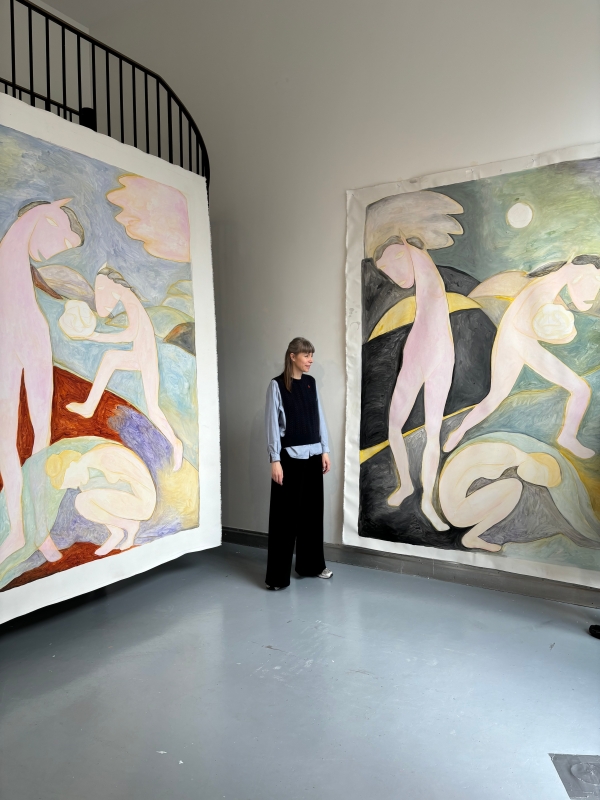 Die Künstlerin
                                          Sarah Bogner steht in einem hohen Ausstellungsraum zwischen zwei großformatigen Gemälden. Die Gemälde zeigen stilisierte,
                                          pastellfarbene Figuren mit angedeuteten Flügeln, fallweise mit Pferdeähnlichen Köpfen in traumartigen Landschaften. Die Künslterin
                                          lächelt. 