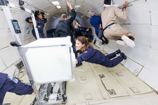 People in blue and beige flight
                                          suits float inside an aircraft cabin during a zero-gravity parabolic flight. Some hold onto the walls or equipment while others
                                          drift freely. In the foreground, one person interacts with a white, cube-shaped piece of equipment as the rest of the group
                                          moves in the background.