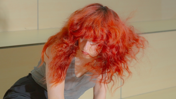 Eine Person mit langen roten Haaren kniet
                                          auf einer hölzernen Plattform.