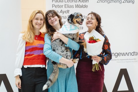 Drei Personen, eine mit Hund auf dem Arm, vor dem Transparent mit der Namensliste
                           aller Absolvent*innen