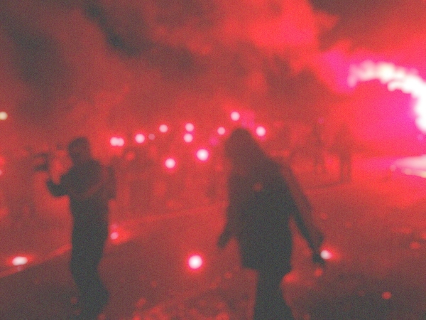 A blurry photograph showing people, red fireworks and smoke.