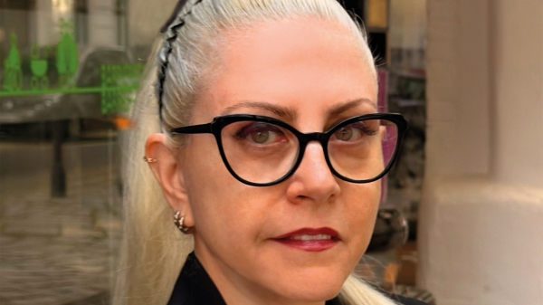 Fotoporträt ovn Basak Senova mit blondem, zurückgebundenem Haar und schwarzer Brille. Sie
                                          trägt ein schwarzes Sakko und eine Halskette und blickt direkt in die Kamera. Im Hintergrund ist ein Ausstellungsraum mit
                                          Glasflächen und Kunstwerken zu sehen.