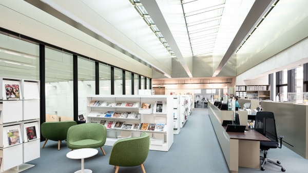 Innenansicht der Universitätsbibliothek
                                          in der Vorderen Zollamtsstraße 7: heller Lesebereich mit modernen Regalen, grünen Sesseln, Zeitschriftenauslage und Informationstheke
                                          unter einem Glasdach.