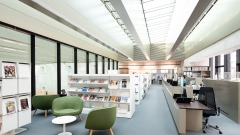 Innenansicht der Universitätsbibliothek in der Vorderen Zollamtsstraße 7: heller Lesebereich mit modernen Regalen, grünen
                                          Sesseln, Zeitschriftenauslage und Informationstheke unter einem Glasdach.