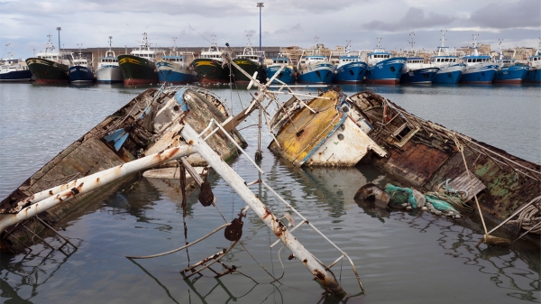 Plakat
                                          zur Ausstellung „Soil & Water: Mediterranean Crossing“ mit einem Foto von versunkenen, beschädigten Fischerbooten im Vordergrund
                                          und einer Reihe intakter Boote im Hafen im Hintergrund. Das Foto stammt aus der Serie Hotspot Mediterraneo (2025) von Francesco
                                          Bellina. Courtesy of the artist.
