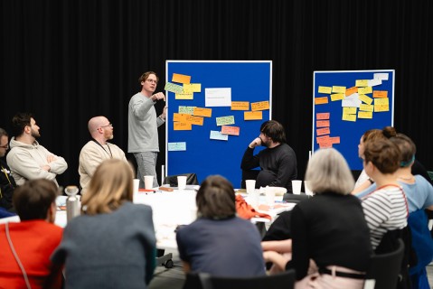 A group of people is sitting at tables working on group projects; a man is standing by a blue bulletin board with Post-ist